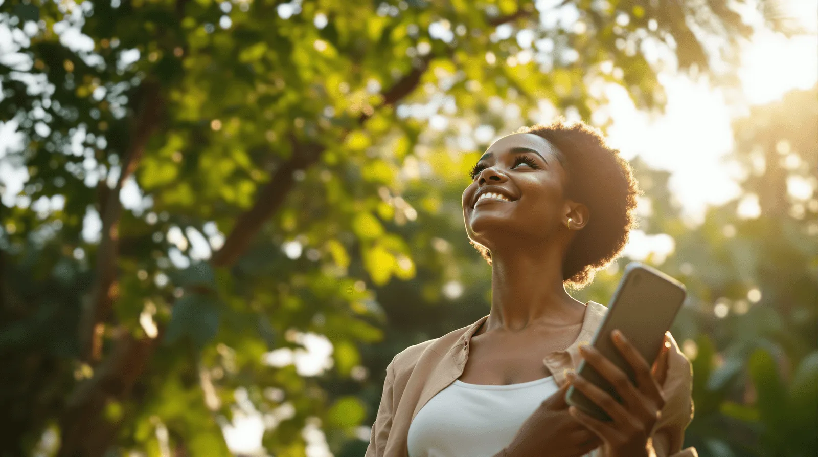Smiling woman holding a phone and looking up at sunlit trees