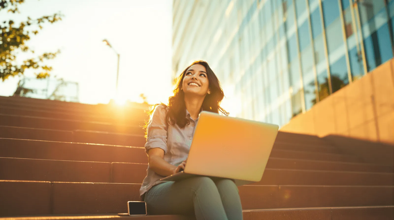 Woman with a laptop smiling on outdoor steps in front of an office building at sunset