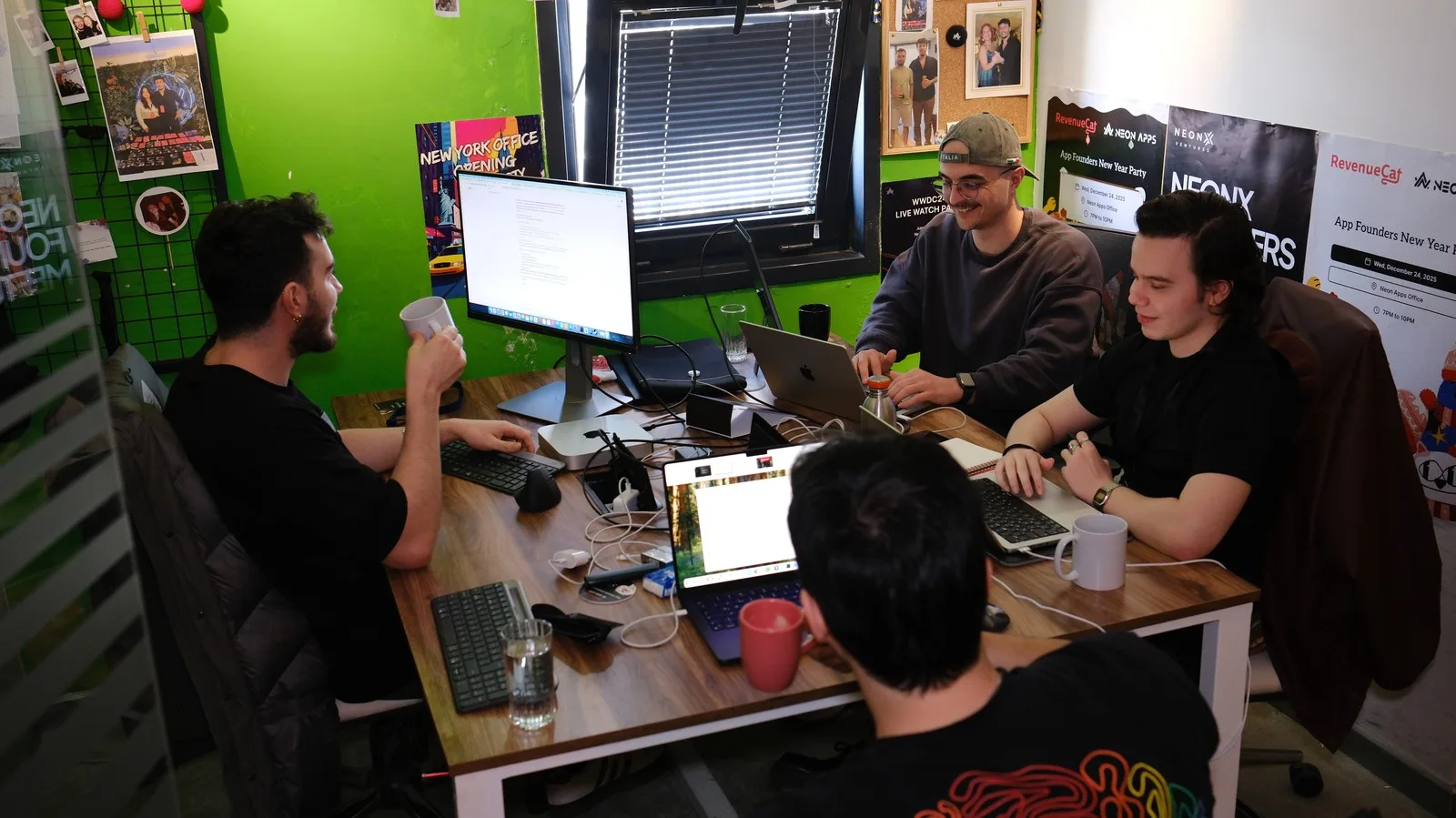 Four colleagues at a shared desk under RevenueCat and Neon Apps event posters