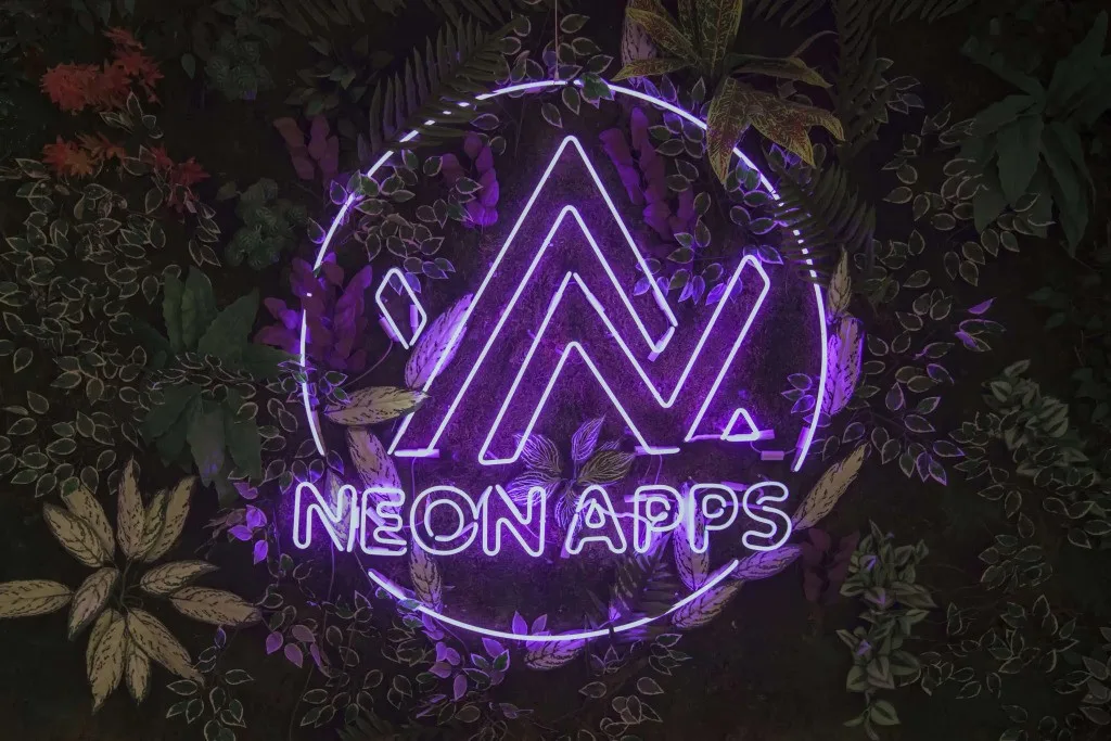 Neon Apps logo displayed as a purple neon sign surrounded by lush green plants, representing innovation in mobile app development and digital technology.